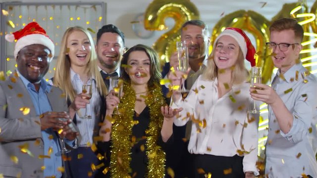 Medium shot of happy businesswomen and businessmen holding glasses of champagne and laughing while posing at New Years party in office as golden confetti falling on them from above