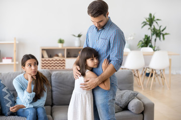 Unhappy diverse family in living room at home. Upset young father embraces sad small daughter,...