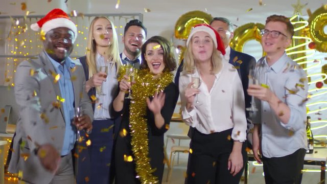 Happy office workers wearing tinsel and Santa hats posing with glasses of champagne at New Years party at work as golden confetti raining on them from above. Number 2019 balloons in background