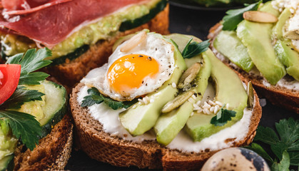 Whole grain bread sandwiches with prosciutto, avocado, cucumber, tomatoes, herbs and seeds in plate on black stone background
