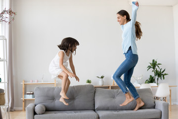 Full length positive mother and little daughter having fun jumping together on sofa in living room...