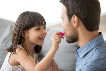 Close up focus on funny girl holding pink lipstick in hand. Cheerful preschool adorable daughter apply cosmetic on father lips. Family have fun sitting on couch at home enjoy time together on weekend