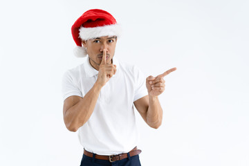 Man wearing Santa hat, pointing aside and making silence gesture. Asian guy touching mouth with forefinger and looking at camera. Christmas surprise concept. Isolated front view on white background.