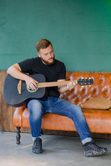 Redhead bearded male with black acoustic guitar play and sitting on the brown leather couch