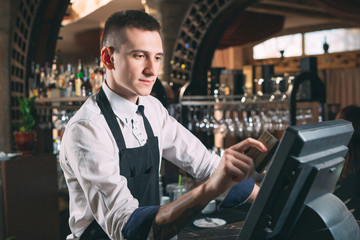 small business, people and service concept - happy man or waiter in apron at counter with cashbox working at bar or coffee shop