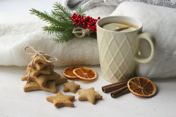 Obraz na płótnie Canvas A Cup of tea with lemon on the table close-up surrounded by Christmas decorations and homemade cakes. Star shaped gingerbread, cinnamon sticks and dried oranges on white background.