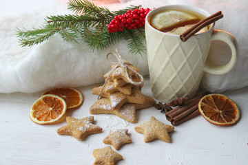 A Cup of tea with lemon on the table close-up surrounded by Christmas decorations and homemade cakes. Star shaped gingerbread, cinnamon sticks and dried oranges on white background.
