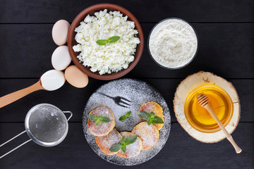 Cottage cheese pancakes and ingredients for their preparation on dark background. Syrniki with honey and mint on plate. Ingredients for cooking: flour, cottage cheese, eggs, sour cream, honey