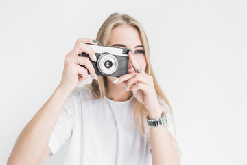 Portrait of a young stylish blonde girl using and taking pictures on an old vintage camera on a white background