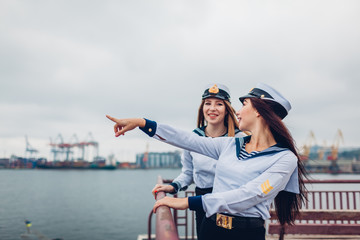 Two college women students of Marine academy walking by sea wearing uniform. Friends pointing into distance on pier
