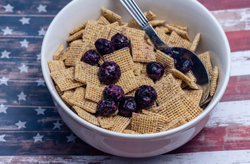 Breakfast cereal in white bowl with frozen blueberries and USA flag background