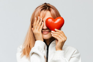 Happy cute young woman with strawberry blonde hair hold red heart (Valentine day symbol),covering her right eye with her palm, closeup, isolated on light background.Love, happiness concept
