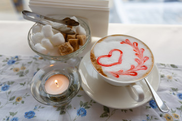 A Cup of hot cappuccino with a pattern of a red heart on the table with a tablecloth in a flower in a coffee shop. Near sugar bowl with sugar and a burning candle. Close-up and blur.