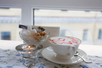 A Cup of hot cappuccino with a pattern of a red heart on the table with a tablecloth in a flower in a coffee shop. Near sugar bowl with sugar and a burning candle. Close-up and blur.