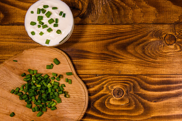 Glass bowl with sour cream and cutting board with chopped green onion on wooden table. Top view