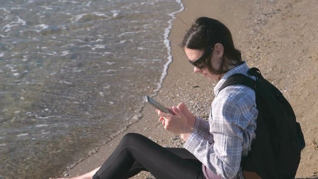 Traveler girl with a backpack sitting on a sandy sea beach and typing a message on mobile phone.