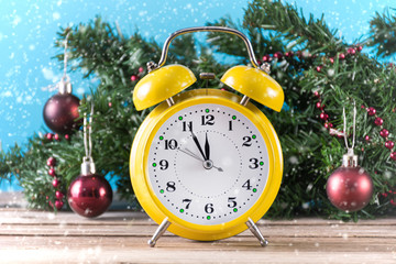 Yellow retro clock on wooden desk and fir Christmas tree with red ball decoration in background. It's snowing at winter day. Christmas and New Year time concept