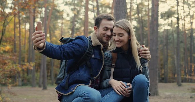 Cheerful young Caucasian couple of tourists sitting in the middle of the forest and taking selfie photo on the smartphone camera. Portrait shot. Outdoors.