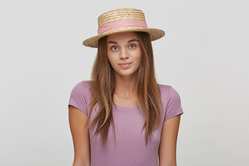 Portrait of beautiful diligent brunette woman in a straw hat with a pink ribbon, looks shy calm, shows tenderness, wears lavender t-shirt, over white background