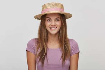 Funny cheerful glad young girl in a straw hat with a pink ribbon, looks playful, bit a lower lip, dressed lavender t-shirt, over white background