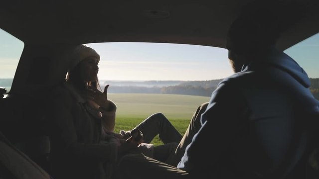 Personal perspective of couple lying in car's trunk, mountain landscape and admiring sunrise . Lovers alking and drinking hot coffee against backdrop of forest panorama in fog . View from inside
