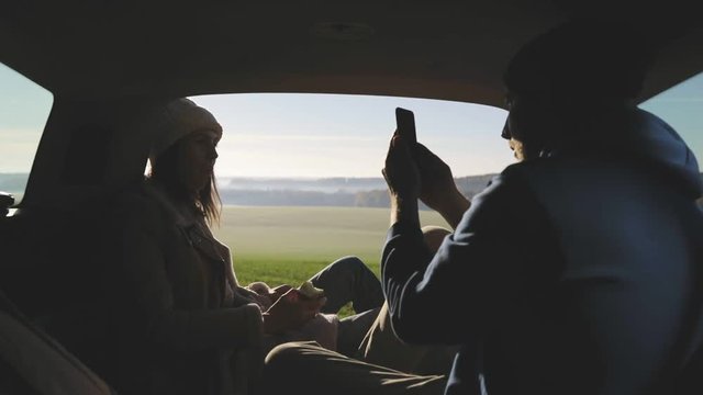 Personal perspective of couple lying in car's trunk, mountain landscape and admiring sunrise . Lovers alking and drinking hot coffee against backdrop of forest panorama in fog . View from inside