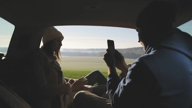 Personal perspective of couple lying in car's trunk, mountain landscape and admiring sunrise . Lovers alking and drinking hot coffee against backdrop of forest panorama in fog . View from inside