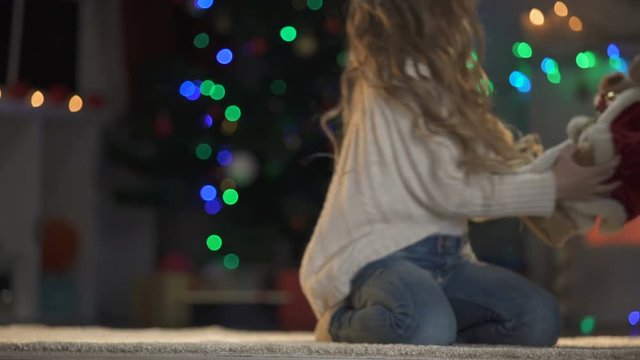 Cute girl playing and hugging teddy bear in Santa outfit, X-mas tree glowing