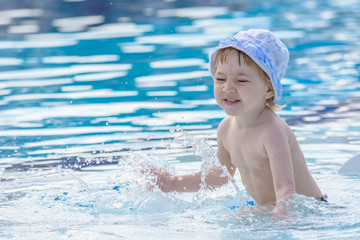 Fototapeta na wymiar Small child in hat disperses waves in the blue pool.