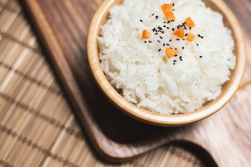 Top view of white rice in wooden bowl with sesame seeds and carrot on wood board