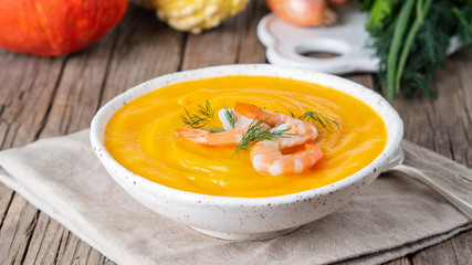 Delicious yellow pumpkin cream soup with shrimp on dark gray old rustic wooden table, side view, long format banner