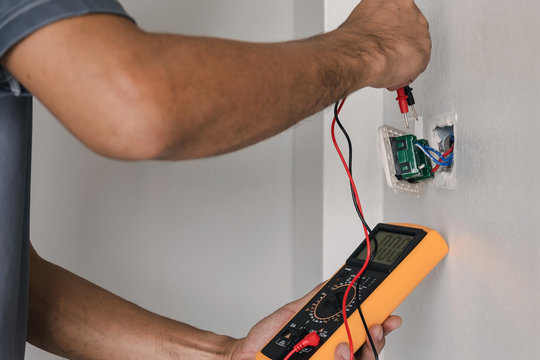 Electrician is using a digital meter to measure the voltage at the power outlet in on the wall.