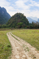 Fototapeta na wymiar Dry footpath between fields leading to Pha Poak, a small limestone hill near Vang Vieng, Vientiane Province, Laos, on a sunny day.