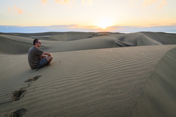 Fototapeta na wymiar Man sitting and relaxing on sand dunes by the sunrise, in Maspalomas on Gran Canaria.