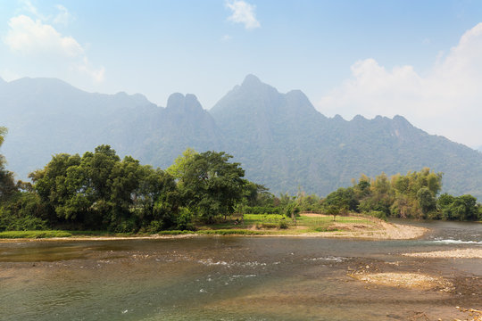 Scenic view of the Nam Song River at low tide, pasture and limestone mountains near Vang Vieng, Vientiane Province, Laos, on a sunny day.