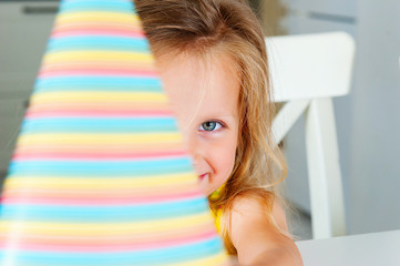 Little cute european blonde girl with blue eyes holding a colorful party hat in front of her face. Close shot