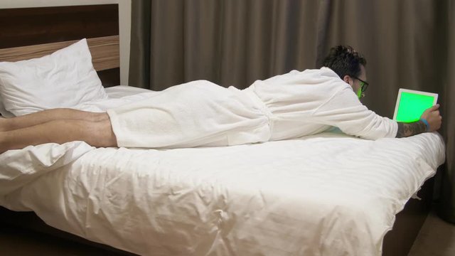 Man looking at data on digital tablet. Man in hotel lies on bed and using touchpad. Businessman working on tablet computer in bad