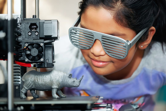 Cute girl with cool 3d printed shutter shades is watching her 3d printer as it prints her 3d model of a rhinoceros.