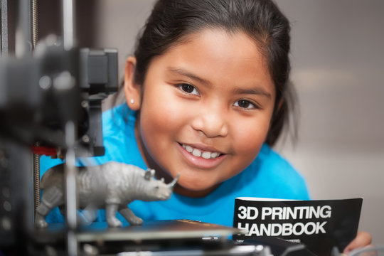 A happy latino girl is proud of her 3d printed toy and is holding a 3d printing handbook as part of a STEM summer camp.