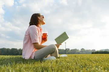 Adult woman resting in the park, sitting on the grass with book and summer refreshment drink, golden hour