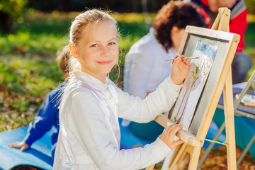 Cute little girl school holding brush painting colorful picture with easel outdoor in park. Open air activity for school age children concept. - obrazy, fototapety, plakaty