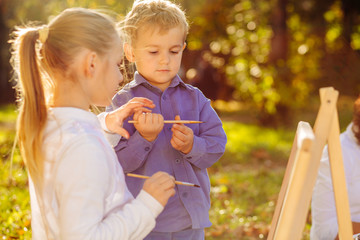 Cute little blond girl school and child boy are holding brushes painting picture with easel outdoor in park. Open air activity for school age children concept.