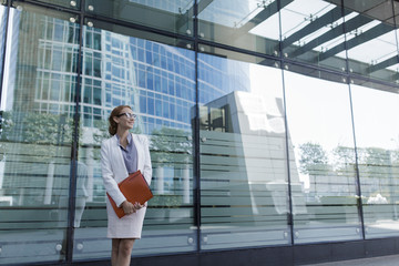 Obraz premium The young business woman in gray office dress holds in hand folder with documents. Businesswoman standing next to the business center