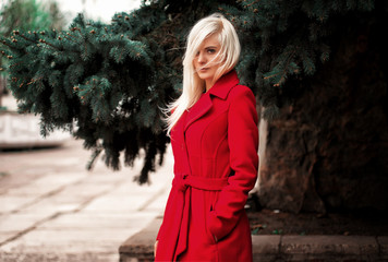 portrait of a blonde woman wearing red coat, near spruce looking in camera