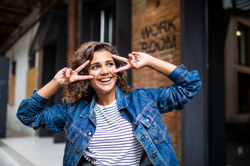 Young latin curly girl wearing a jeans jacket with peace gesture standing on a brick background on a street.