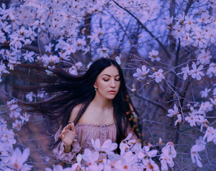 wonderful attractive dark-haired lady with eyes closed stands in the garden of blooming magnolias. hair flies up with the wind, amazing photo in cold shades of violet filter, art processing photos