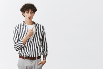 Indoor shot of impressed and amazed jewish curly-haired guy with moustache and tattoos dropping jaw and gasping from interest and excitement pointing at upper right corner intrigued