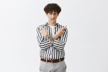 Fototapeta premium Help me make decision. Weak and insecure concerned stylish caucasian guy with curly hairstyle and moustache crossing hands on chest pointing left and right biting lip being unsure which way right