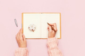 Female hands in pink knitted sweater written with a pen in open clean notebook and cotton on pink...
