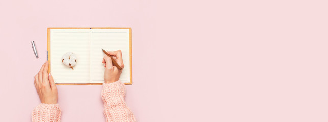 Female hands in pink knitted sweater written with a pen in open clean notebook and cotton on pink pastel table top view flat lay Fashion blogger working desk Cotton flowers Lifestyle gentle background